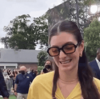 A Woman Wearing Sunglasses And A Yellow Shirt Is Smiling In Front Of A Crowd Of People . GIF