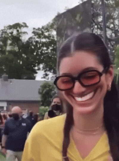 A Woman Wearing Sunglasses And A Yellow Shirt Is Smiling While Standing In Front Of A Crowd . GIF