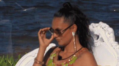 A Woman Wearing Sunglasses And Earrings Is Sitting On A Chair Near The Water . GIF