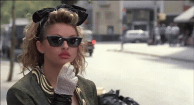 A Woman Wearing Sunglasses And Gloves Is Standing On A Street . GIF
