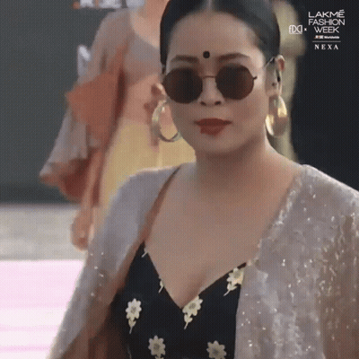 A Woman Wearing Sunglasses And Hoop Earrings Is Walking Down A Runway At Lakme Fashion Week GIF
