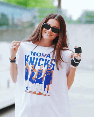 A Woman Wearing Sunglasses Holds Up A Nova Knicks Shirt GIF