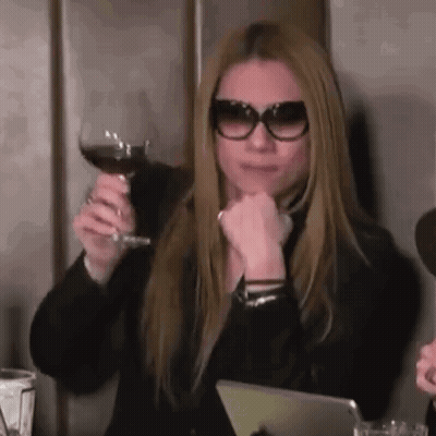 A Woman Wearing Sunglasses Is Sitting At A Table Holding A Glass Of Red Wine . GIF
