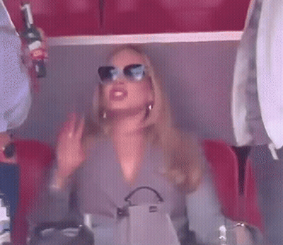 A Woman Wearing Sunglasses Is Sitting In A Stadium Holding A Bottle Of Wine And A Purse . GIF