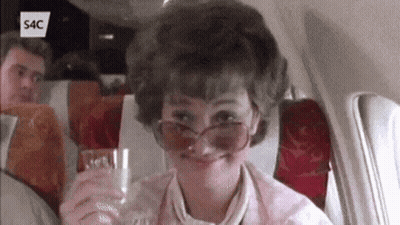 A Woman Wearing Sunglasses Is Sitting On An Airplane Holding A Glass GIF