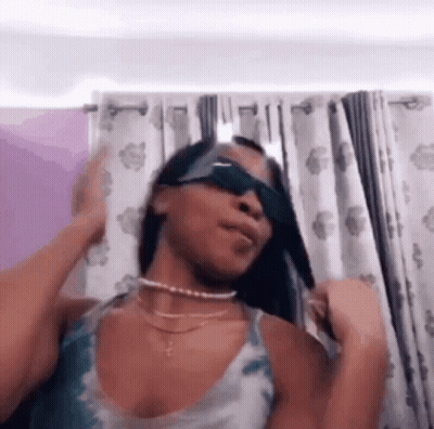 A Woman Wearing Sunglasses Is Standing In Front Of A Curtain . GIF