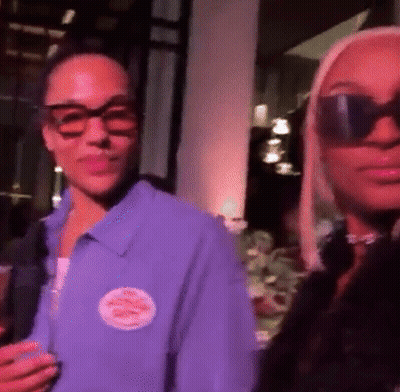 A Woman Wearing Sunglasses Is Standing Next To A Man Wearing A Purple Shirt . GIF