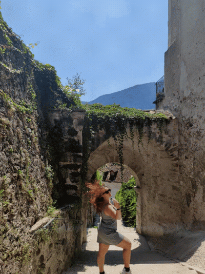 A Woman Wearing Sunglasses Stands In A Stone Archway GIF
