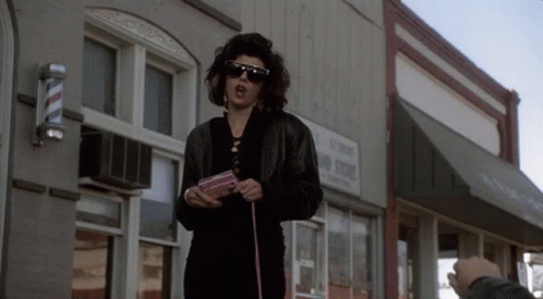A Woman Wearing Sunglasses Stands In Front Of A Building With A Sign That Says No Stop GIF
