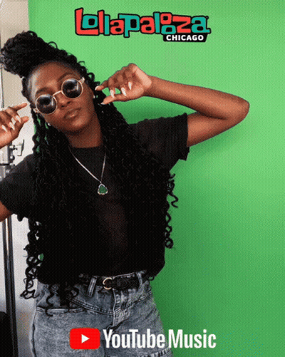 A Woman Wearing Sunglasses Stands In Front Of A Green Screen That Says ' Lolapaloza Chicago ' On It GIF