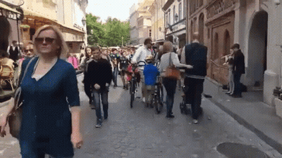 A Woman Wearing Sunglasses Walks Down A Busy Street With People Walking And Riding Bikes GIF