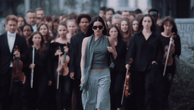 A Woman Wearing Sunglasses Walks In Front Of A Large Group Of Musicians GIF