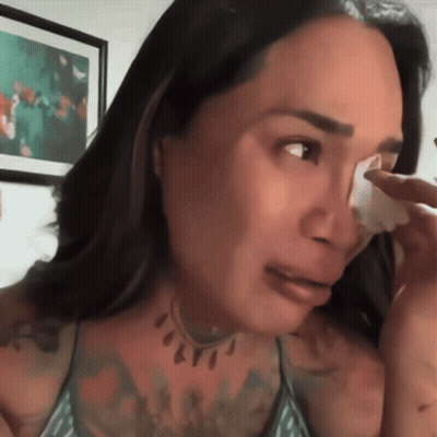 A Woman Wipes Her Face With A Tissue In Front Of A Picture GIF