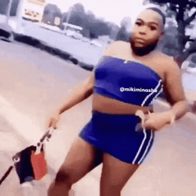 A Woman With A Beard Is Wearing A Blue Top And Shorts And Holding A Bag . GIF