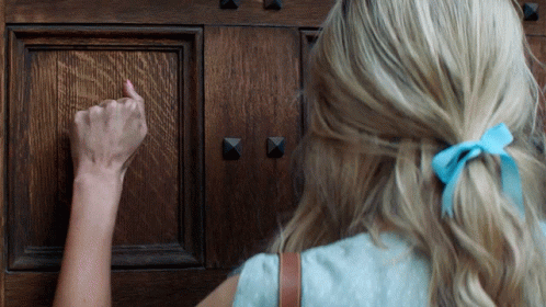 A Woman With A Blue Bow In Her Hair Knocks On A Door GIF