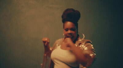 A Woman With A Bun On Her Head Is Wearing Hoop Earrings And A Sequined Jacket GIF