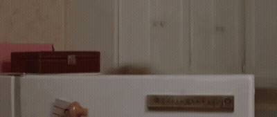 A Woman With A Face Mask On Her Face Is Standing In Front Of A Refrigerator . GIF
