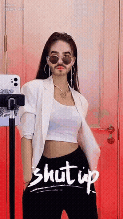 A Woman With A Fake Mustache And Sunglasses Is Standing In Front Of A Red Wall . GIF
