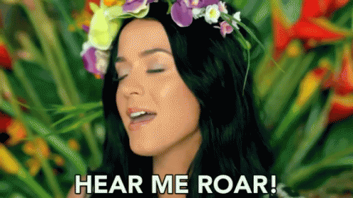A Woman With A Flower Crown On Her Head Is Standing In Front Of Flowers And Says `` Hear Me Roar '' . GIF