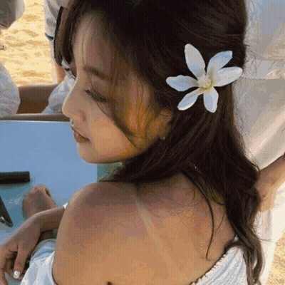 A Woman With A Flower In Her Hair Is Sitting On The Beach GIF