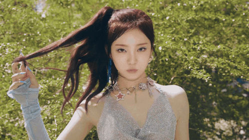 A Woman With A Ponytail And A Star Necklace Stands In Front Of Trees GIF