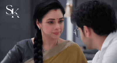 A Woman With A Red Dot On Her Forehead Looks At A Man With Glasses In Front Of A Sign That Says Sk GIF