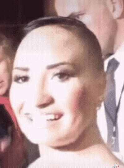 A Woman With A Shaved Head Is Smiling In Front Of A Man In A Suit And Tie . GIF