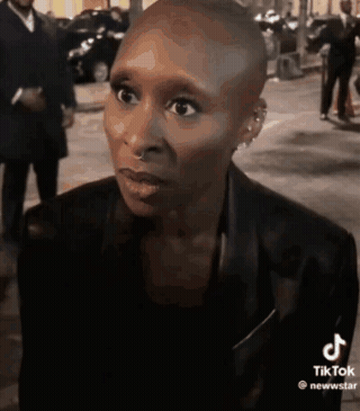 A Woman With A Shaved Head Is Wearing A Black Jacket And Earrings And Looking At The Camera . GIF