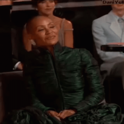 A Woman With A Shaved Head Is Wearing A Green Dress While Sitting In A Audience . GIF