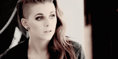 A Woman With A Shaved Head Is Wearing Earrings And A Black Jacket . GIF