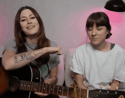 A Woman With A Tattoo On Her Arm Is Playing A Guitar Next To Another Woman GIF