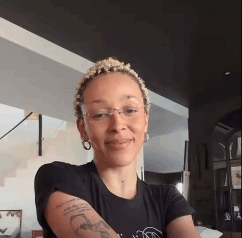 A Woman With A Tattoo On Her Arm Is Wearing Glasses And A Black Shirt GIF