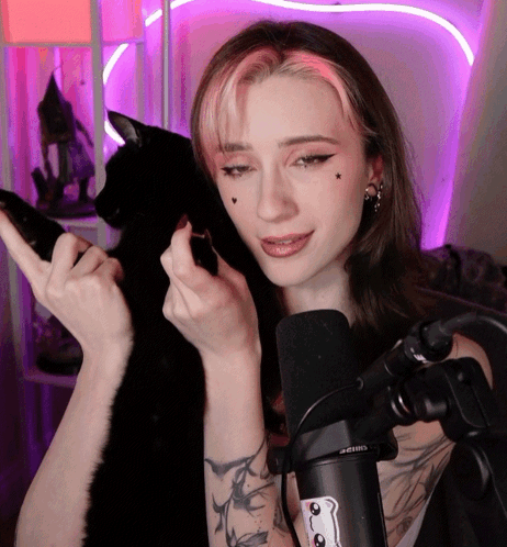 A Woman With A Tattoo On Her Arm Stands In Front Of A Microphone And A Black Cat GIF
