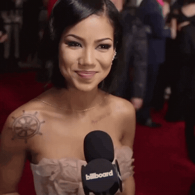 A Woman With A Tattoo On Her Shoulder Is Smiling And Holding A Billboard Microphone GIF