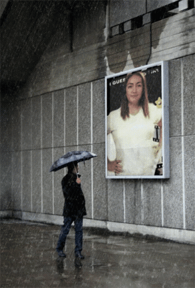 A Woman With An Umbrella Stands In Front Of A Billboard That Says I Guess GIF