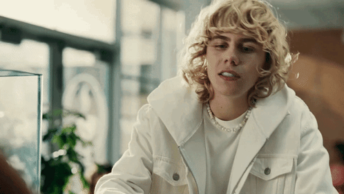 A Woman With Blonde Curly Hair Wearing A White Jacket And A Pearl Necklace GIF