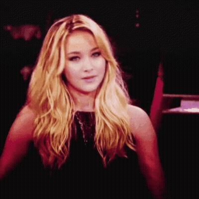 A Woman With Blonde Hair And A Black Dress Looks At The Camera GIF