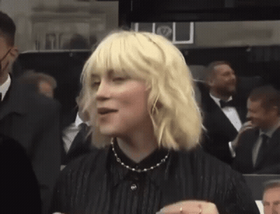 A Woman With Blonde Hair And A Black Shirt Is Standing In A Crowd . GIF