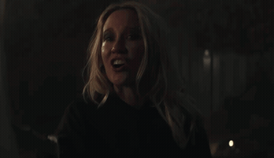 A Woman With Blonde Hair And A Black Shirt Smiles In The Dark GIF