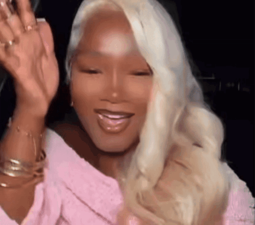 A Woman With Blonde Hair And A Pink Shirt Is Waving At The Camera . GIF