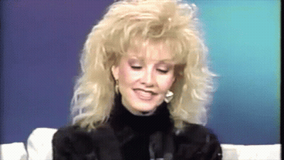 A Woman With Blonde Hair And A Turtleneck Is Smiling GIF