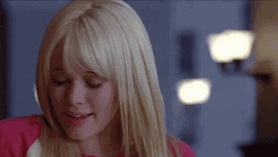 A Woman With Blonde Hair And Bangs Is Smiling While Wearing A Pink Shirt . GIF