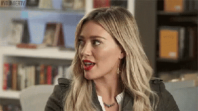 A Woman With Blonde Hair And Red Lips Is Sitting In Front Of A Bookshelf And Making A Funny Face . GIF