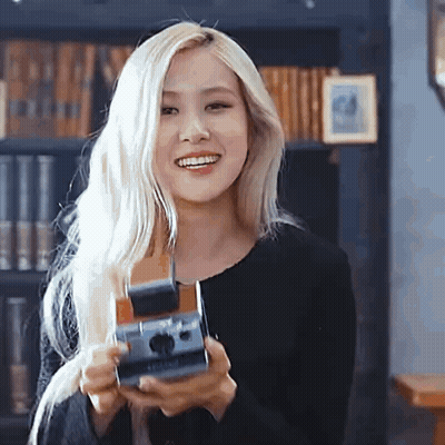 A Woman With Blonde Hair Is Holding A Camera In Her Hands And Smiling . GIF