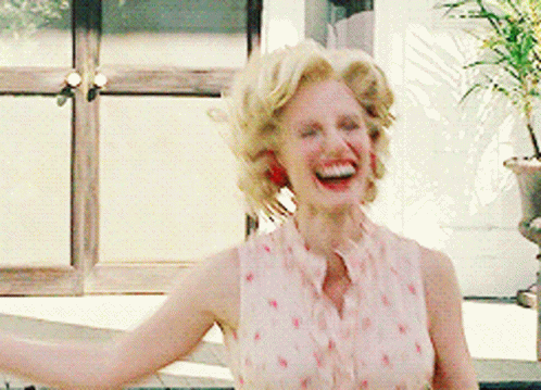 A Woman With Blonde Hair Is Laughing With Her Arms Outstretched In Front Of A Window GIF