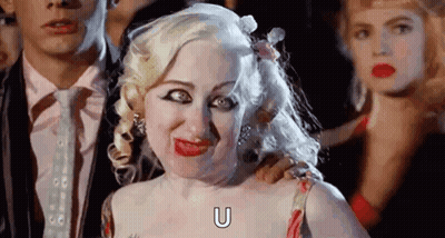 A Woman With Blonde Hair Is Making A Funny Face While Standing Next To A Man In A Suit . GIF