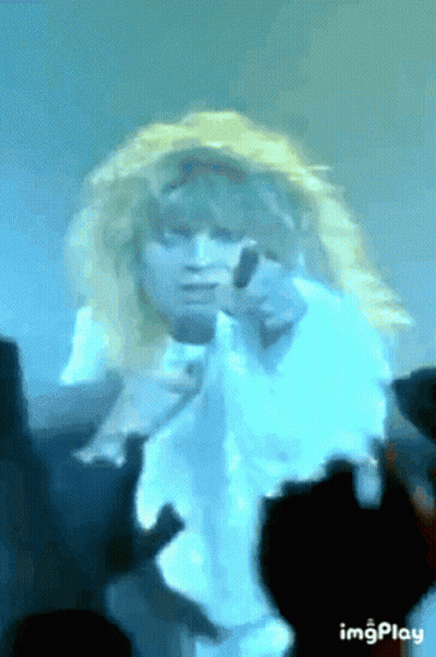 A Woman With Blonde Hair Is Singing Into A Microphone While A Crowd Watches . GIF