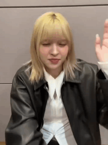 A Woman With Blonde Hair Wearing A Black Leather Jacket And A White Shirt Is Sitting In Front Of A Wall . GIF