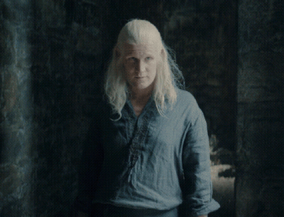 A Woman With Blonde Hair Wearing A Blue Shirt Stands In A Dark Room GIF