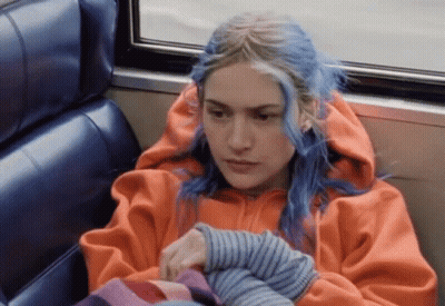 A Woman With Blue Hair And An Orange Hoodie Is Sitting On A Bus GIF